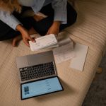 Vista de cima de uma mulher organizando finanças na cama, com notebook e papéis ao lado
