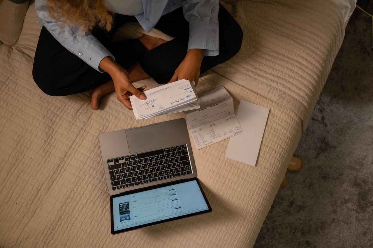 Vista de cima de uma mulher organizando finanças na cama, com notebook e papéis ao lado