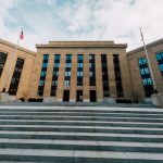 Fachada do Federal Reserve em Washington, vista de baixo, com bandeiras dos Estados Unidos e escadaria sob céu nublado.