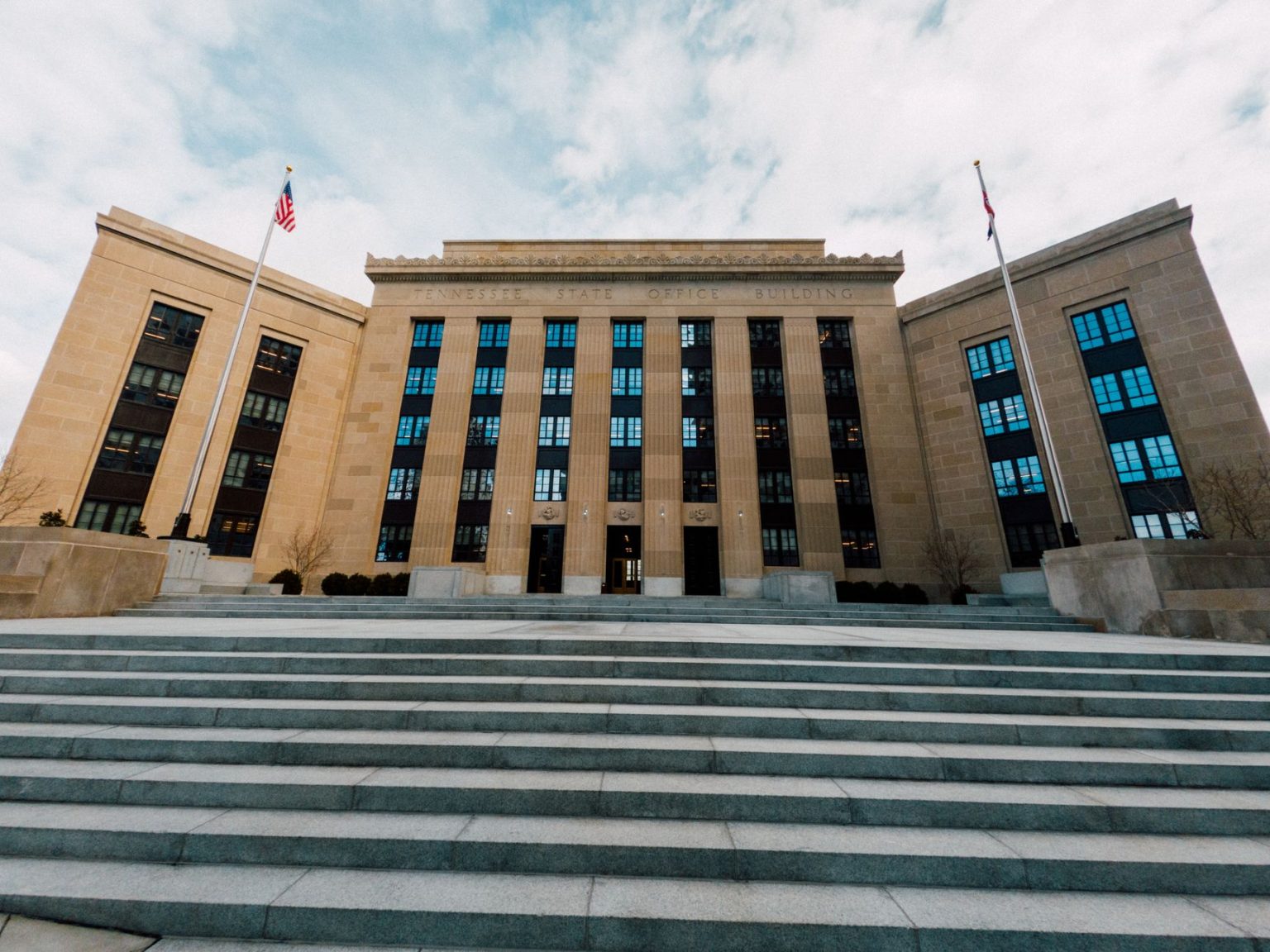 Fachada do Federal Reserve em Washington, vista de baixo, com bandeiras dos Estados Unidos e escadaria sob céu nublado.