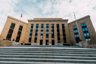 Fachada do Federal Reserve em Washington, vista de baixo, com bandeiras dos Estados Unidos e escadaria sob céu nublado.