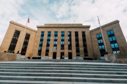 Fachada do Federal Reserve em Washington, vista de baixo, com bandeiras dos Estados Unidos e escadaria sob céu nublado.