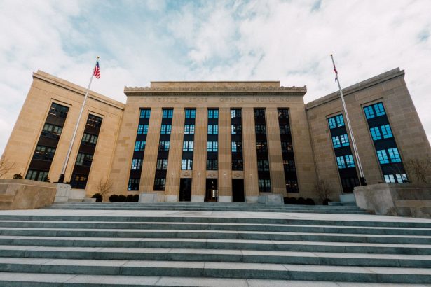 Fachada do Federal Reserve em Washington, vista de baixo, com bandeiras dos Estados Unidos e escadaria sob céu nublado.
