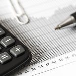 Calculadora, caneta e documentos financeiros sobre uma mesa, representando cálculos orçamentários e análise fiscal
